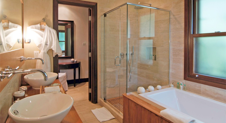 Spacious modern bathroom with a glass shower enclosure, warm wooden tub surround, two vessel sinks, and soft golden lighting, tranquil mood