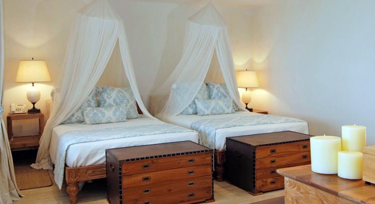 Bedroom with two twin beds under sheer white canopies, blue and white patterned pillows, wooden trunks at the foot of the beds, tranquil tropical mood